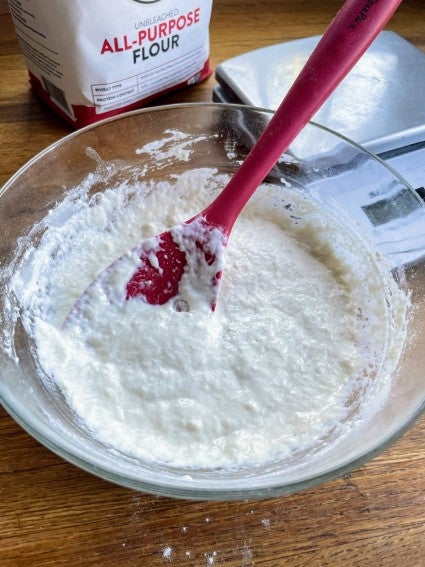 Stirring fed sourdough starter in a clear glass bowl, bag of King Arthur Flour and a digital scale in the background.